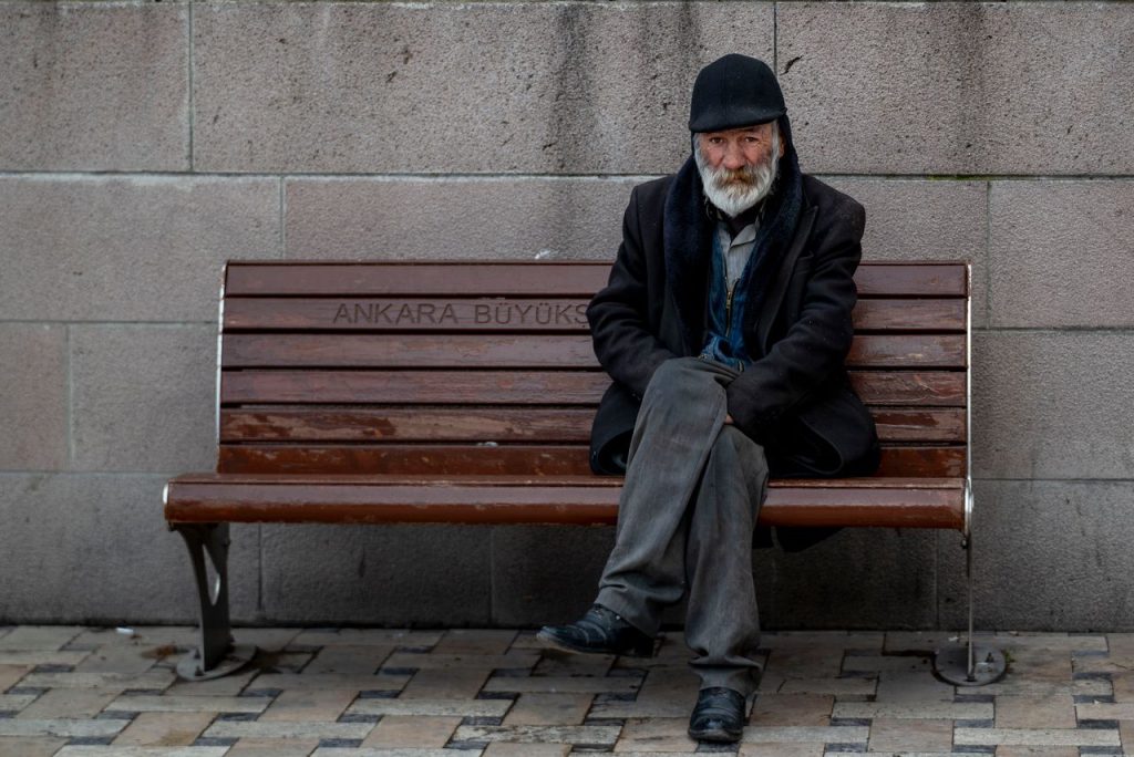 Elderly man with a beard sitting on a bench in Ankara, wearing a coat and cap.