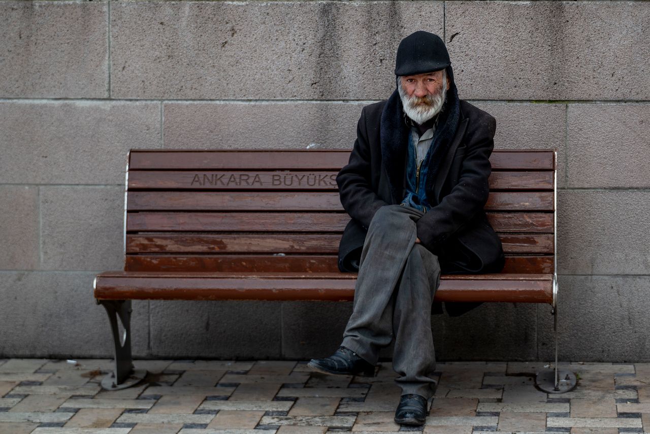 Elderly man with a beard sitting on a bench in Ankara, wearing a coat and cap.